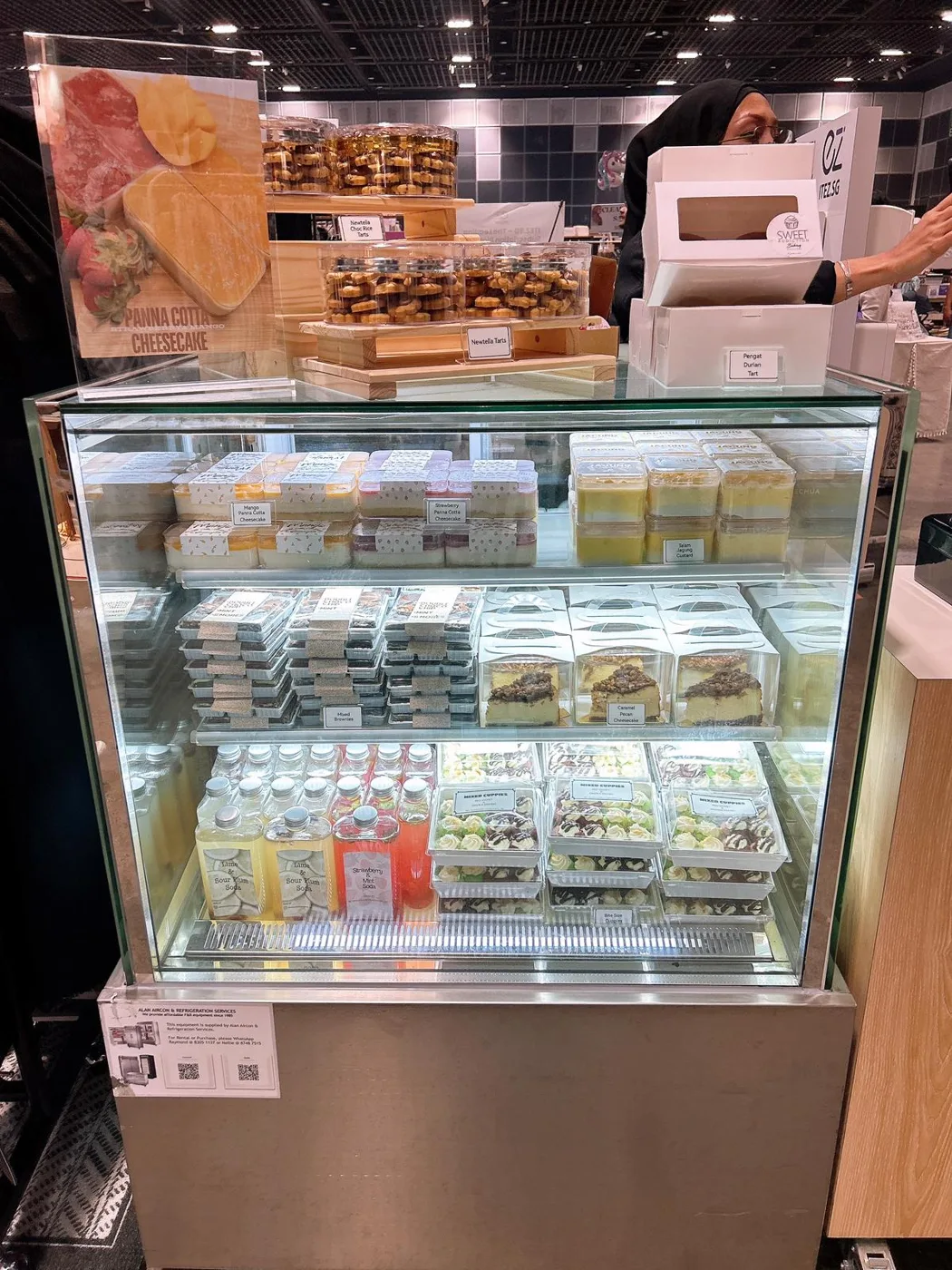 AANRS cake chiller rental Singapore, three shelves of boxed cakes, tarts and bottled drinks inside a glass display at an event stall